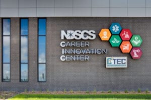 Brick exterior with the sign that says "NDSCS Career Innovation Center" and light-up visual graphics. Three tall skinny windows to the right. Grass and shrubbery below.