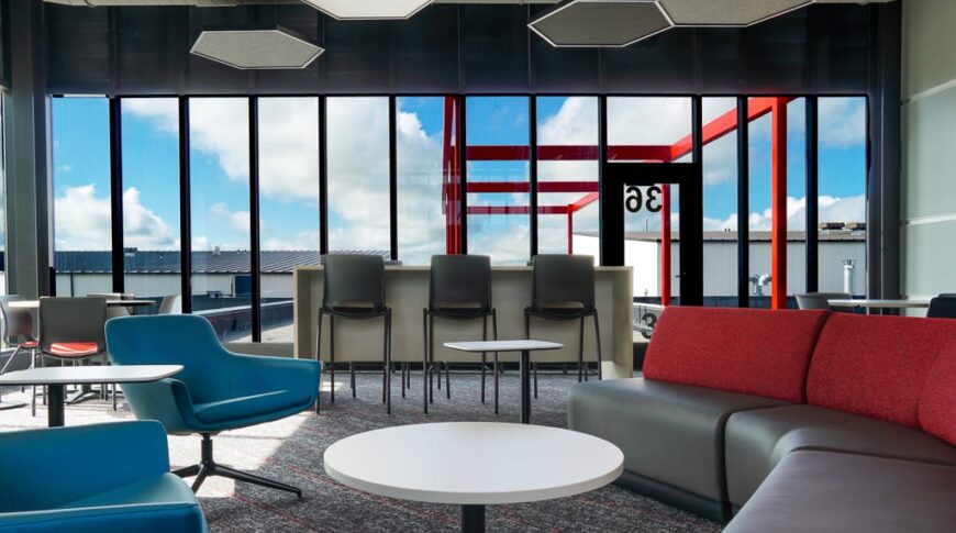 Hangout Den on second story of NDSCS Career Innovation Center with curved lounge couch, accent chairs, and bar-top station behind, all overlooking bright blue skies through the floor to ceiling windows.