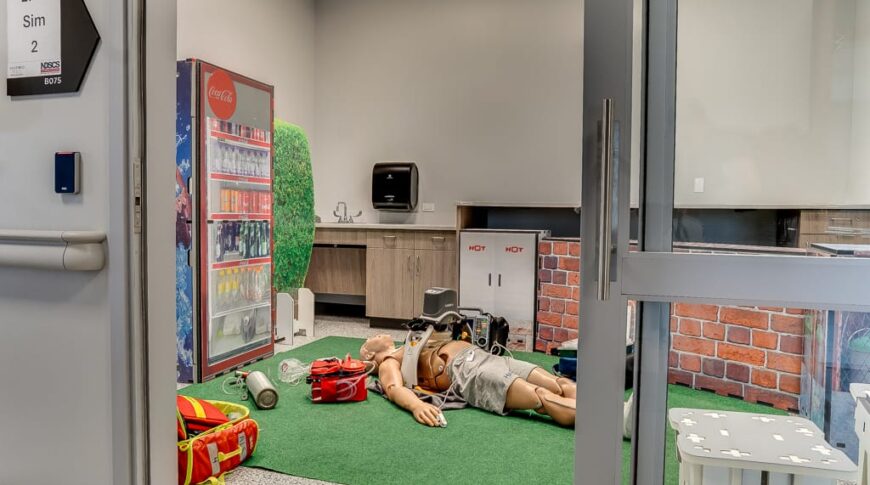 Simulation of medical emergency with manikin on turf grass surrounded by faux barbecue area with vending machine in the background.
