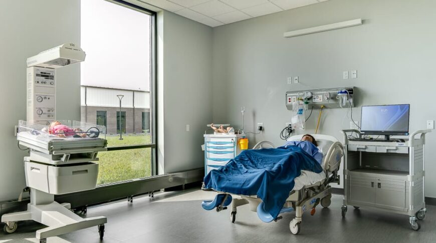 Maternity care room simulator with mother medical manikin on hospital bed and baby manikin under heating lamp station.
