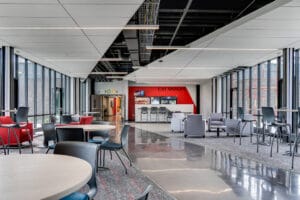 View of entire commons area at NDSCS Career Innovation Center with tables and chairs and seating areas mixed throughout, and a vending station to the back wall. Both right and left sides of the room featuring floor to ceiling windows, with modern partly exposed ceiling above.
