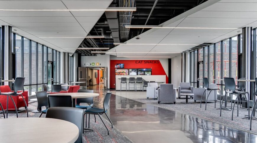 View of entire commons area at NDSCS Career Innovation Center with tables and chairs and seating areas mixed throughout, and a vending station to the back wall. Both right and left sides of the room featuring floor to ceiling windows, with modern partly exposed ceiling above.