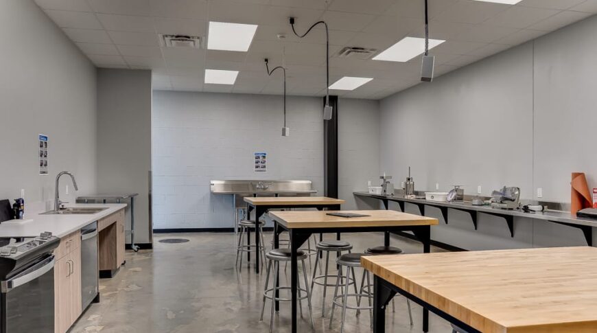 Meat processing classroom complete with kitchen workstation along the left wall, processing equipment on the right wall, and bar-height tables and stools in the center.