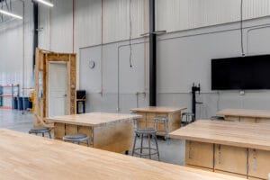 Light wood-toned workstations and stools of the construction workshop with a replica door and window construct for demonstrations.