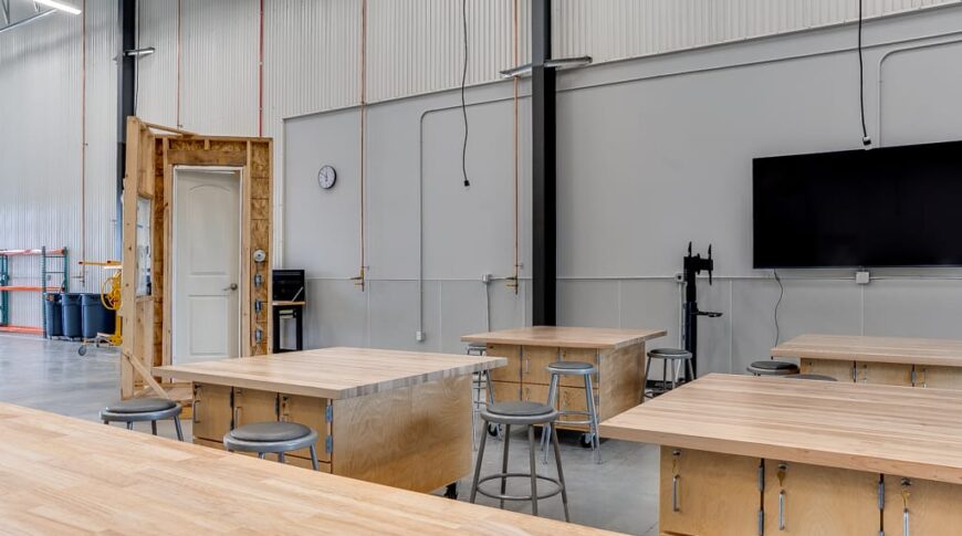 Light wood-toned workstations and stools of the construction workshop with a replica door and window construct for demonstrations.