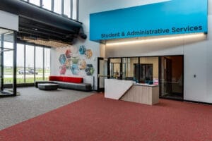Reception area with blue awning that reads "Student & Administrative Services" with a desk in front. Loung area to the back alongside a wall of multi-story floor to ceiling windows.