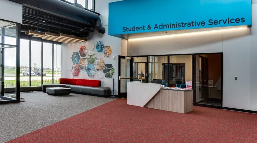 Reception area with blue awning that reads "Student & Administrative Services" with a desk in front. Loung area to the back alongside a wall of multi-story floor to ceiling windows.