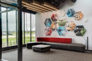 Entryway lounge at NDSCS Career Innovation Center with floor to ceiling windows overlooking the landscaping, modern modular gray and red couch, brightly-colored hexagon graphic on wall and modern plank overhead lighting.