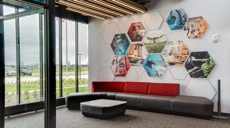 Entryway lounge at NDSCS Career Innovation Center with floor to ceiling windows overlooking the landscaping, modern modular gray and red couch, brightly-colored hexagon graphic on wall and modern plank overhead lighting.