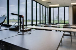 Classroom view from the perspective of the teacher's desk overlooking rows of tables and chairs and two walls of floor to ceiling windows.