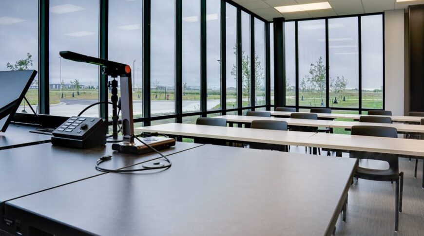 Classroom view from the perspective of the teacher's desk overlooking rows of tables and chairs and two walls of floor to ceiling windows.