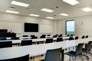 Classroom with teacher's desk and workstation at the front left with dry erase boards and monitor on the front wall, with rows of tables and chairs ahead.