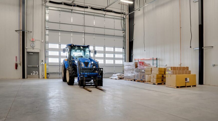 Construction bay with tractor inside and palleted materials to the right wall, with closed garage bay doors behind.