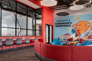 Den area of college with back wall graphic in bright blue and orange hued colors that says "Empowering Tomorrow's Workforce." To the back left a wall of countertop and chair stations for working, and in the center a curved sofa and accent chairs station.