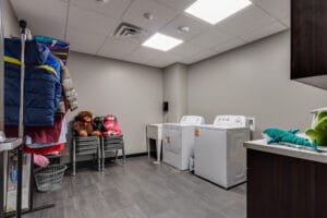 Laundry room with grey-colored walls and laminate flooring, washer and dryer to back left wall, racks of clothing hanging on left wall.