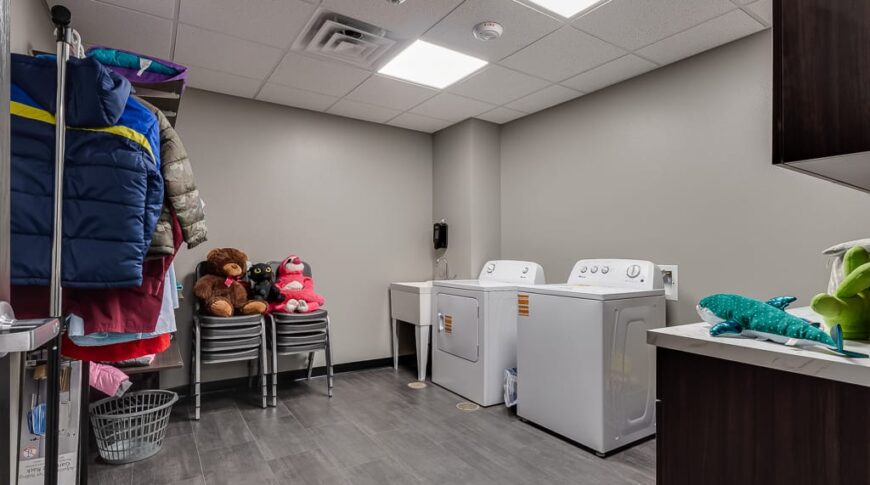 Laundry room with grey-colored walls and laminate flooring, washer and dryer to back left wall, racks of clothing hanging on left wall.