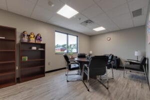 Employee breakroom with light gray colored walls and LVP flooring, a small round table and chairs surrounding, some bookcases to the back left wall besides a window.
