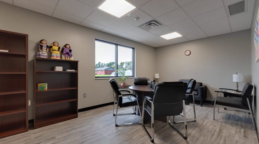 Employee breakroom with light gray colored walls and LVP flooring, a small round table and chairs surrounding, some bookcases to the back left wall besides a window.