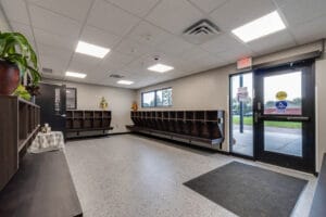 Entryway to Enemy Swim Head Start with large windowed door and sidelight to the right, and rows of cubbies for backpacks and jackets along the three perimeter walls seen in the image.