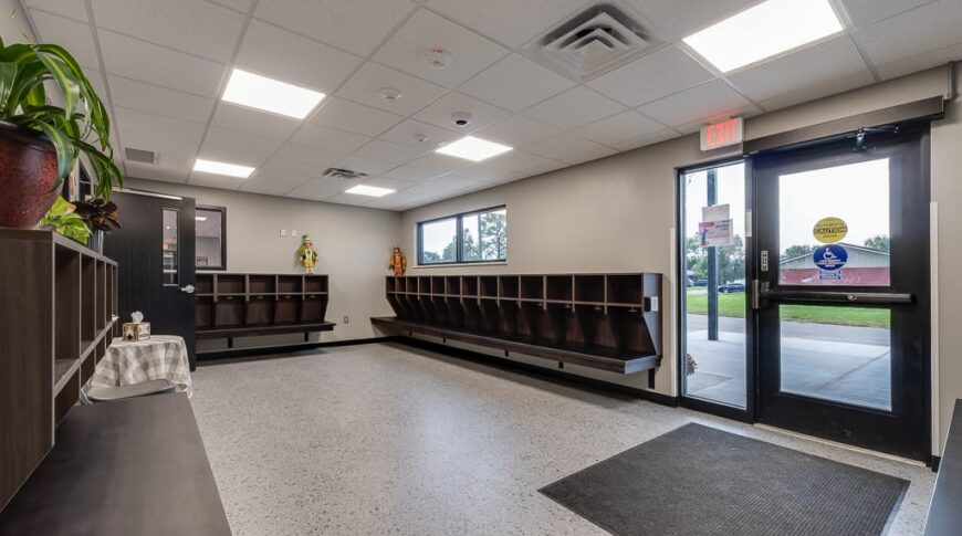 Entryway to Enemy Swim Head Start with large windowed door and sidelight to the right, and rows of cubbies for backpacks and jackets along the three perimeter walls seen in the image.