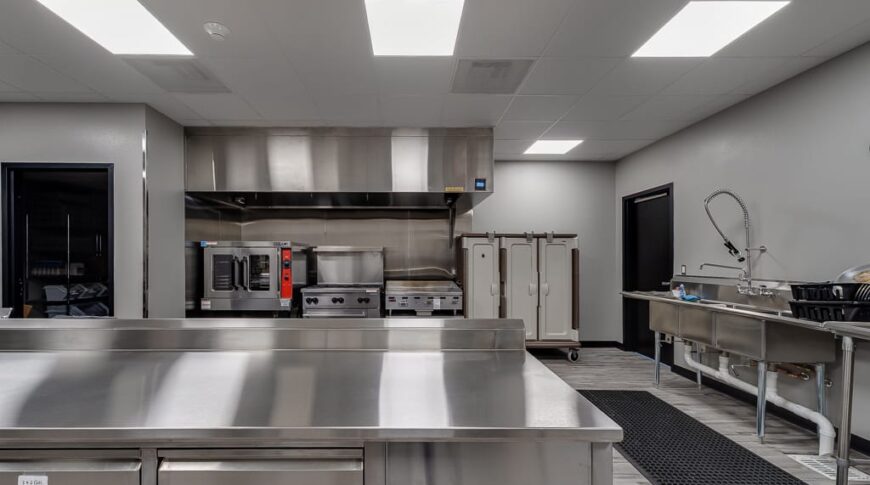 Industrial kitchen at Enemy Swim Head Start overlooking stainless steel island and oven and stove to the far back wall and washing station to the right.