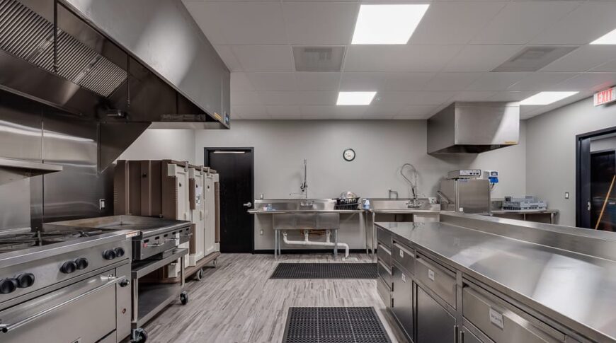 Industrial kitchen with stainless steel island to the right and appliances to the left. Wash station to the back wall.