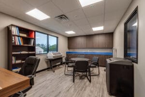 Business center and supply room with desk and chair to the left, bookcase and printer with window on wall to the back left, upper and lower wall of cabinetry on back wall, and right wall a shredder with interior window overlooking classrooms. In the middle of the room is a small round table and four chairs.