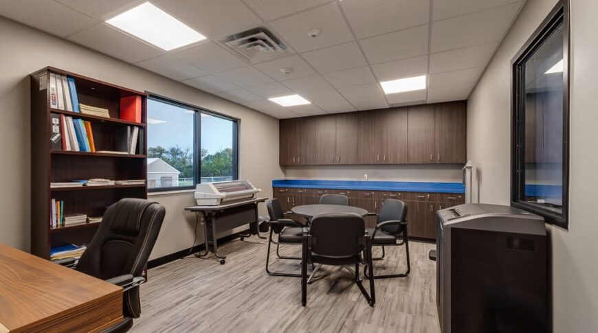 Business center and supply room with desk and chair to the left, bookcase and printer with window on wall to the back left, upper and lower wall of cabinetry on back wall, and right wall a shredder with interior window overlooking classrooms. In the middle of the room is a small round table and four chairs.
