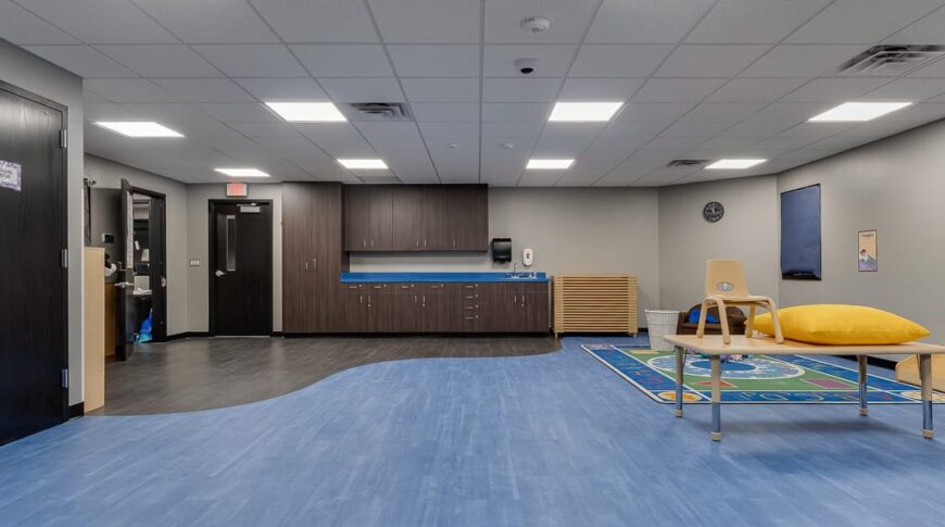 Large kids classroom with blue and gray laminate flooring. Upper and lower cabinets to the back wall and play area to the right.