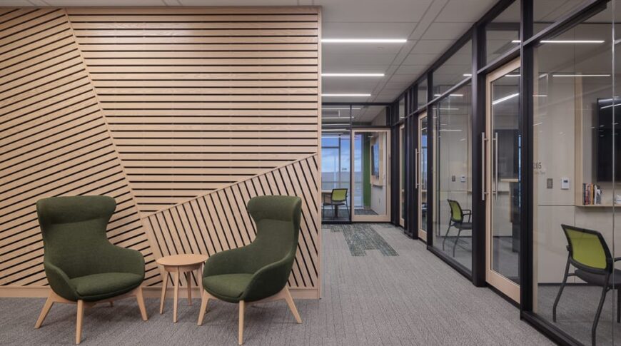 Seating area with two wingback chairs in front of wooden slat accent wall, with row of floor-to-ceiling office windows to the right.