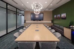 Conference room with accent lighting above long light-wooden table flanked by gray accent chairs. Green painted wall to the right, floor-to-ceiling windows to the left and a wooden accent wall to the far back wall with a TV monitor.