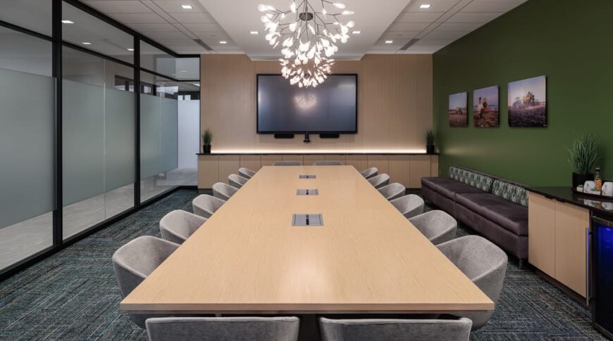 Conference room with accent lighting above long light-wooden table flanked by gray accent chairs. Green painted wall to the right, floor-to-ceiling windows to the left and a wooden accent wall to the far back wall with a TV monitor.