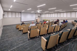 Auditorium with five rows of risers with folding chairs looking to the front podium and drop-down projector screen.
