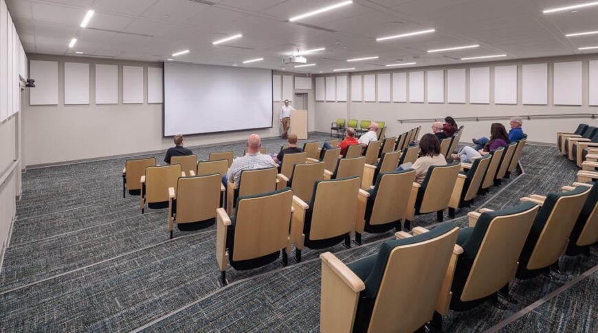 Auditorium with five rows of risers with folding chairs looking to the front podium and drop-down projector screen.
