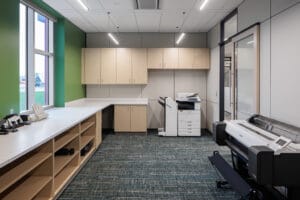Printing station room with green painted wall with window to the left with cabinetry and countertop below. Upper and lower cabinetry and printer to the back wall, and plotter printer to the right wall.