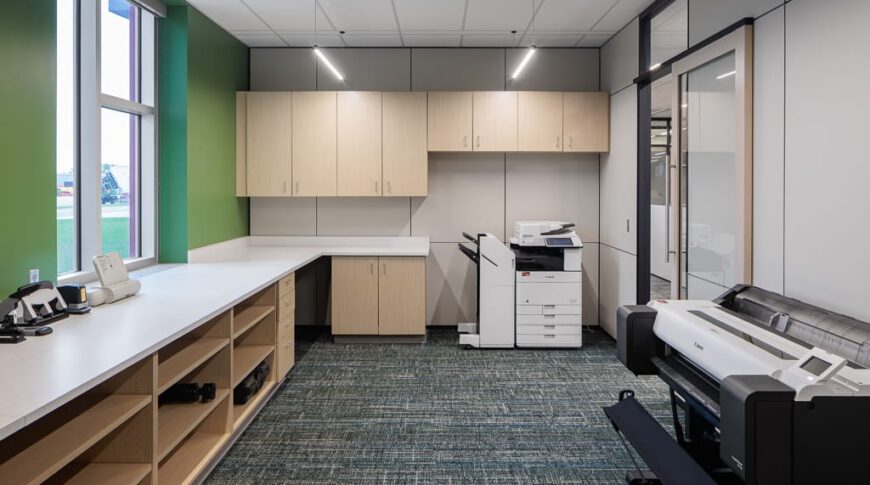 Printing station room with green painted wall with window to the left with cabinetry and countertop below. Upper and lower cabinetry and printer to the back wall, and plotter printer to the right wall.