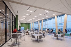 Large, open dining area with bar and regular-height tables and chairs throughout and wall of windows looking outside to the right and wall of windows looking to the interior of the building to the left.