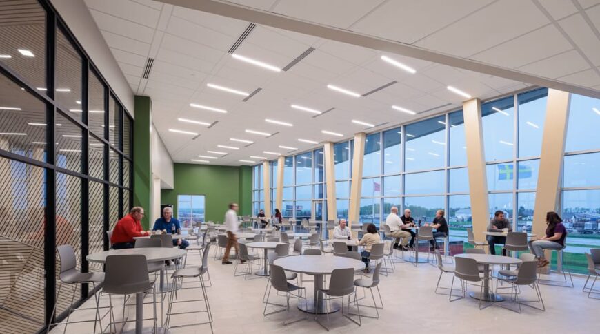 Large, open dining area with bar and regular-height tables and chairs throughout and wall of windows looking outside to the right and wall of windows looking to the interior of the building to the left.