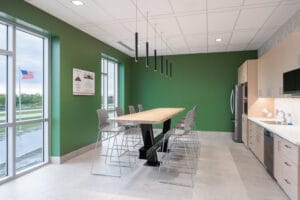 Full kitchen with galley-style row of cabinets, fridge, and dishwasher to the right, bar-height table and chairs in the middle, and green-painted walls with floor-to-ceiling windows the left.