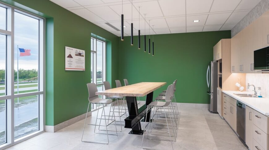 Full kitchen with galley-style row of cabinets, fridge, and dishwasher to the right, bar-height table and chairs in the middle, and green-painted walls with floor-to-ceiling windows the left.