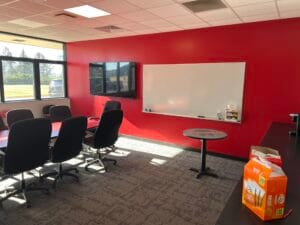 Newly remodeled conference room at NDSCS Blikre Activities Center. Bright red accent wall with dry erase board and tv to the back wall. Windows to the left wall, conference table in the middle, and countertop station to the right.