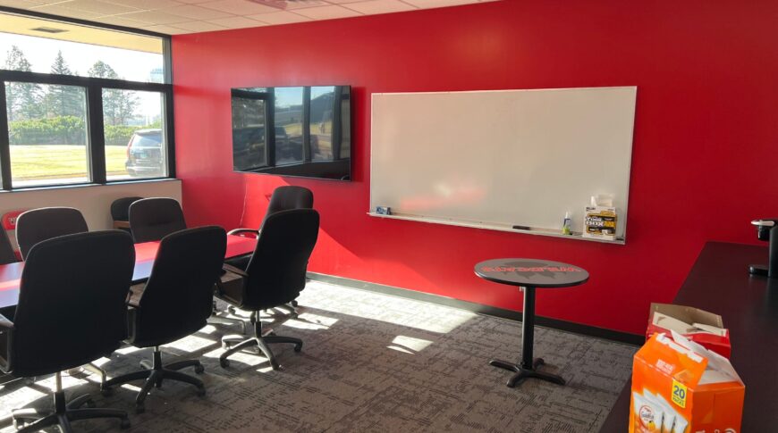 Newly remodeled conference room at NDSCS Blikre Activities Center. Bright red accent wall with dry erase board and tv to the back wall. Windows to the left wall, conference table in the middle, and countertop station to the right.