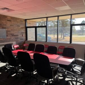 Conference room with red table and black swivel chairs surrounding in the middle of the room. Back wall of windows behind the table and brick wall to the back left.