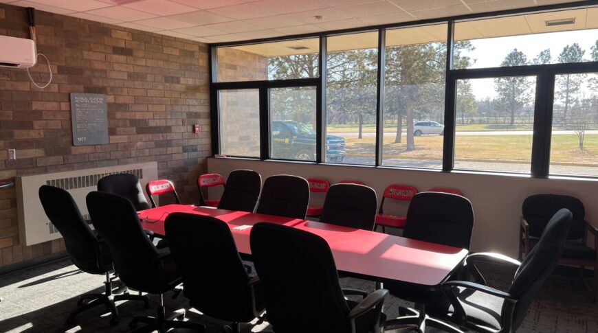 Conference room with red table and black swivel chairs surrounding in the middle of the room. Back wall of windows behind the table and brick wall to the back left.