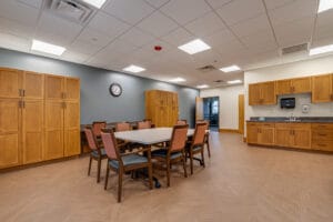 Newly remodeled gathering room at PioneerCare Center in Fergus Falls, with large meeting table and chairs in the center of the room, flanked by a wall of tall cabinetry behind and a kitchenette to the back right.