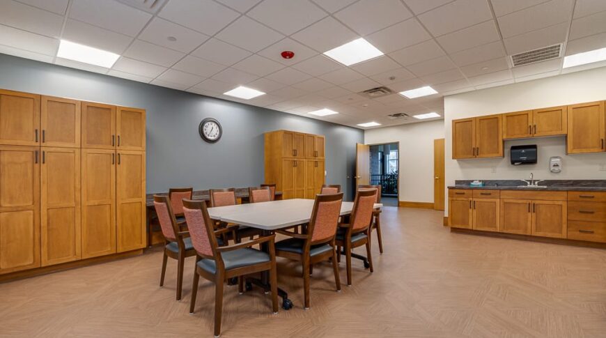 PioneerCare-Low Res-20 Newly remodeled gathering room at PioneerCare Center in Fergus Falls, with large meeting table and chairs in the center of the room, flanked by a wall of tall cabinetry behind and a kitchenette to the back right.