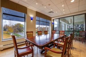 Large floor to ceiling windows replace a previous railing in a nursing home meeting room, that overlooks an interior garden and waterfall. Large table and chairs in middle of the room with exterior windows overlooking grounds behind.