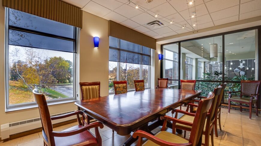 PioneerCare-Low Res-27 Large floor to ceiling windows replace a previous railing in a nursing home meeting room, that overlooks an interior garden and waterfall. Large table and chairs in middle of the room with exterior windows overlooking grounds behind.