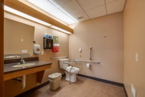 Dimly lit beige bathroom that was newly remodeled for a nursing home facility in Fergus Falls. Accessible sink and vanity to the left with toilet and rails to the back.