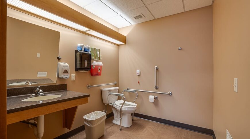 PioneerCare-Low Res-34 Dimly lit beige bathroom that was newly remodeled for a nursing home facility in Fergus Falls. Accessible sink and vanity to the left with toilet and rails to the back.